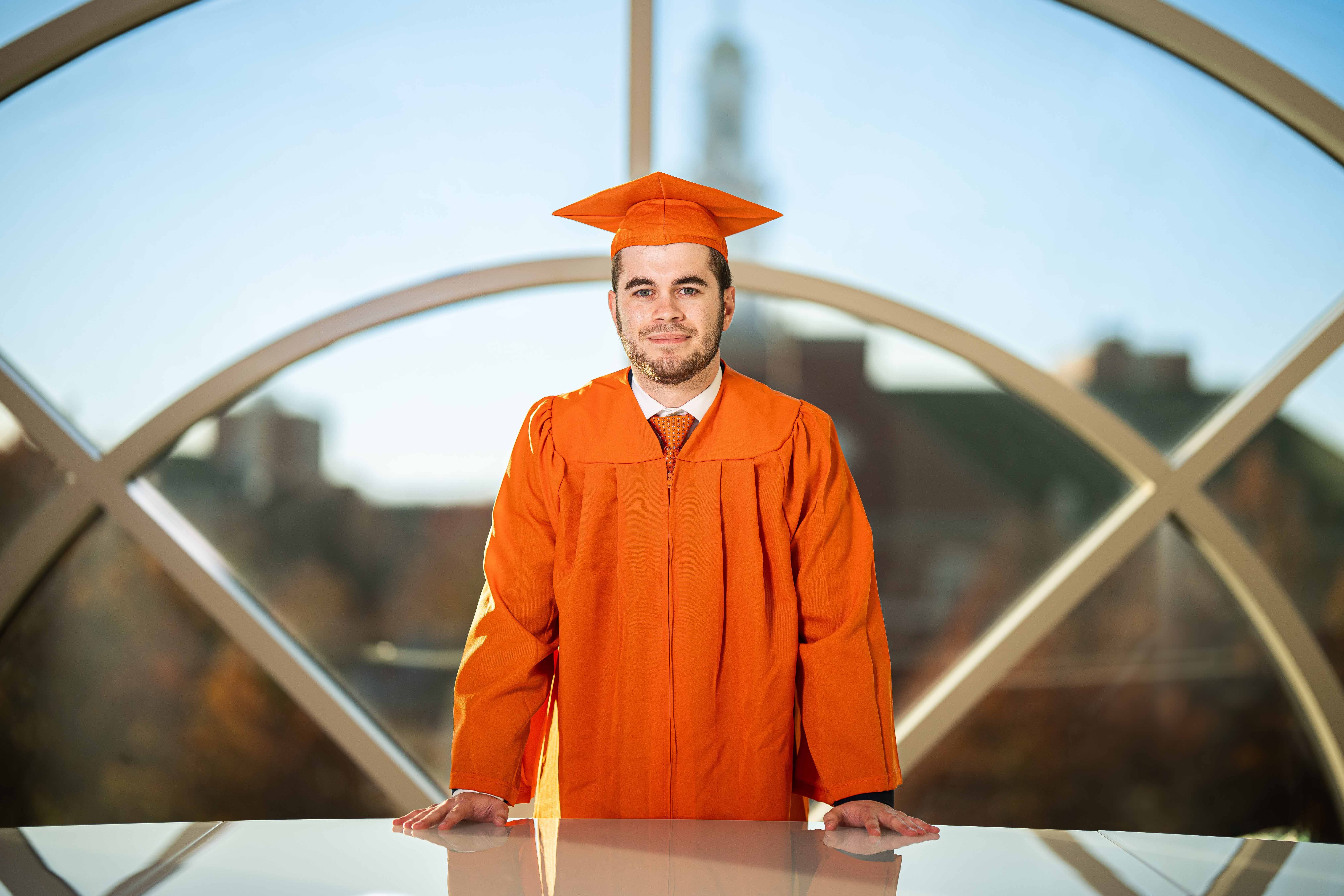 Student wearing an orange cap and gown standing confidentley and leaning on a table in a nice room in the Spears School of Business.