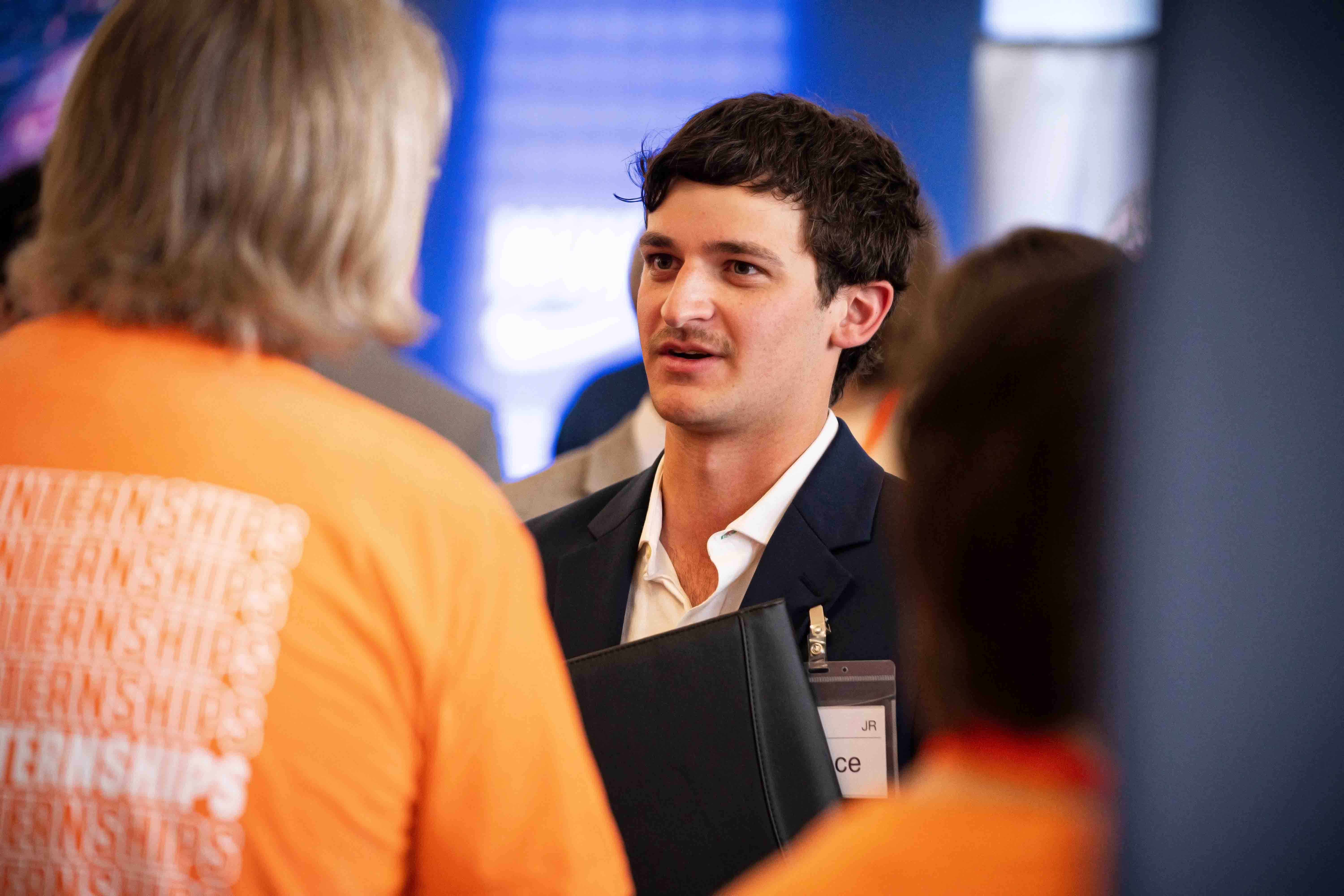 Male student holding portfolio (folder for resumes) and speaking to company at the Career Fair.