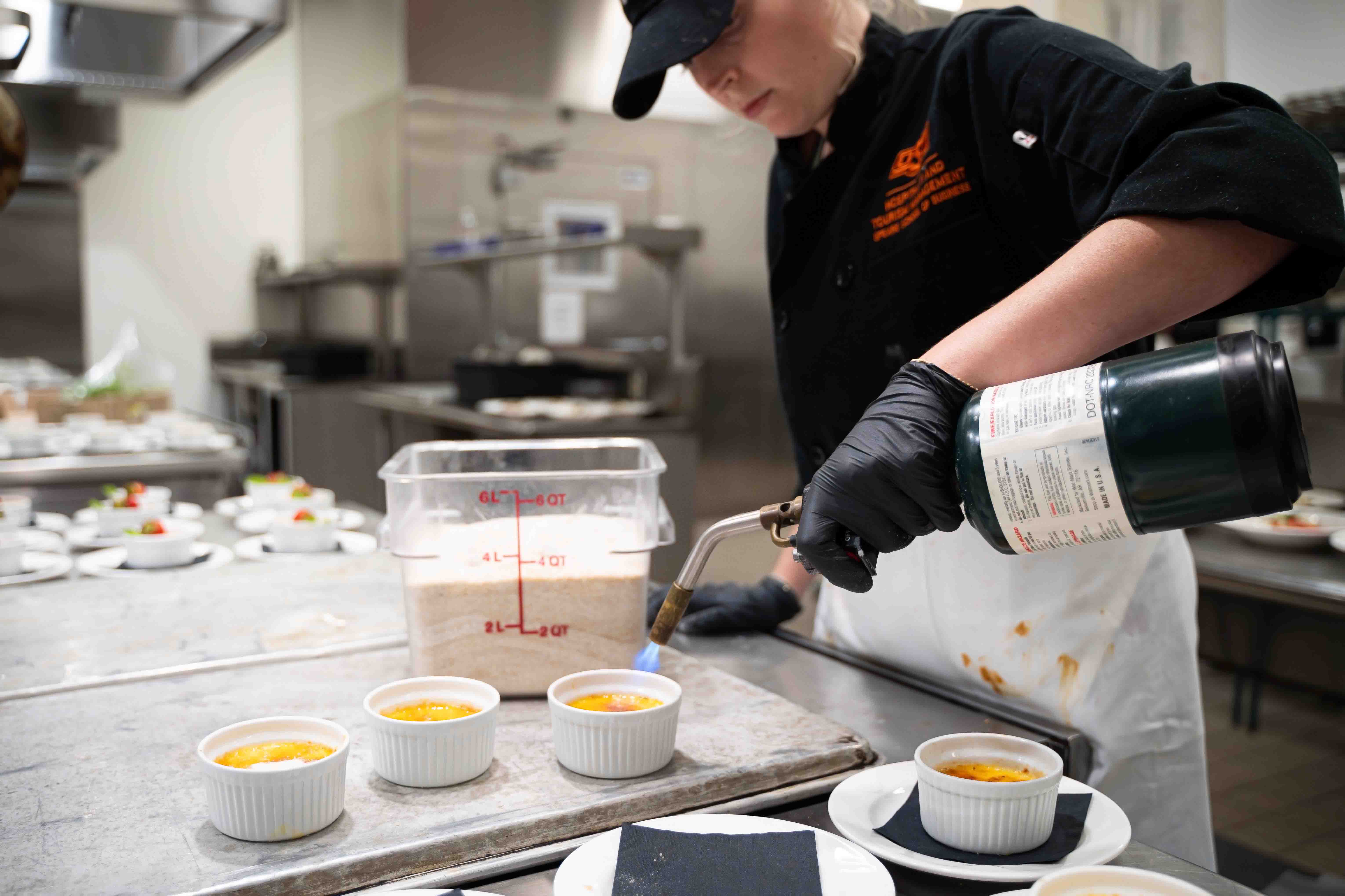 Student holding a kitchen torch carmelizing the sugar on top of a creme brulee. Student is wearing a chef's coat and is inside the industrial kitchen in the HTM building.