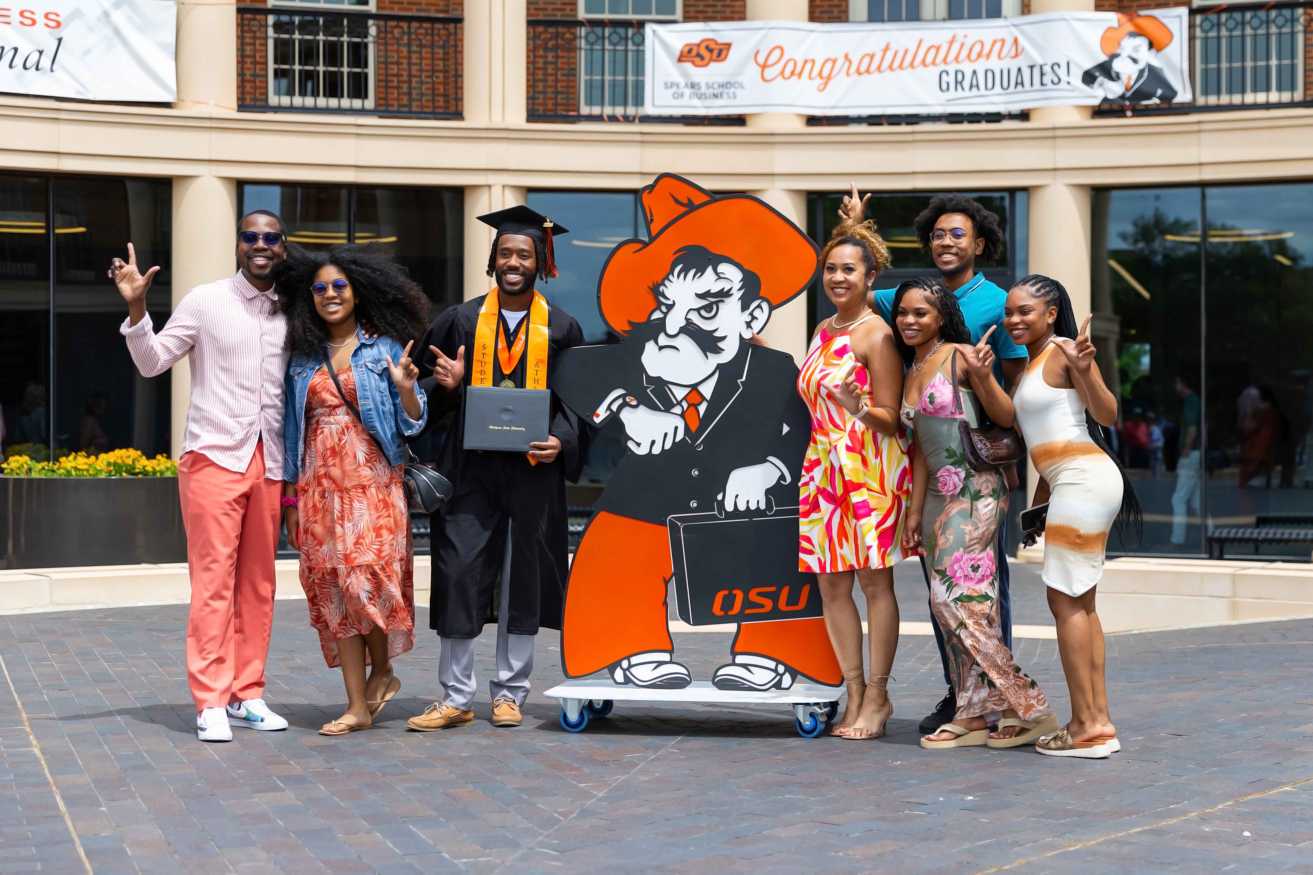 Student standing with family after graduation. Smiling with Business Pete metal mascot outside of the Business Building.