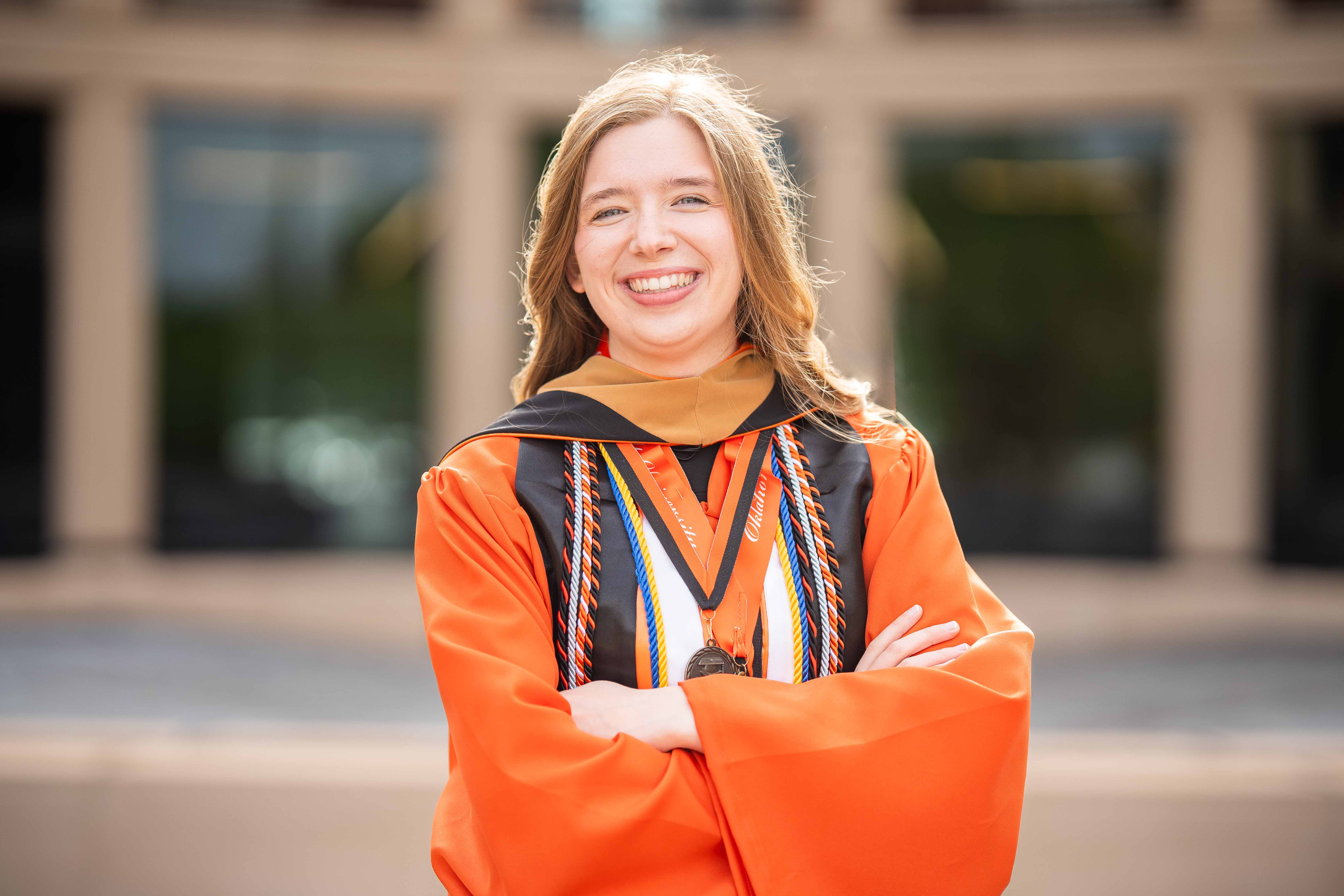 Female graduate wearing an orange graduation gown smiling in front of the Business Building