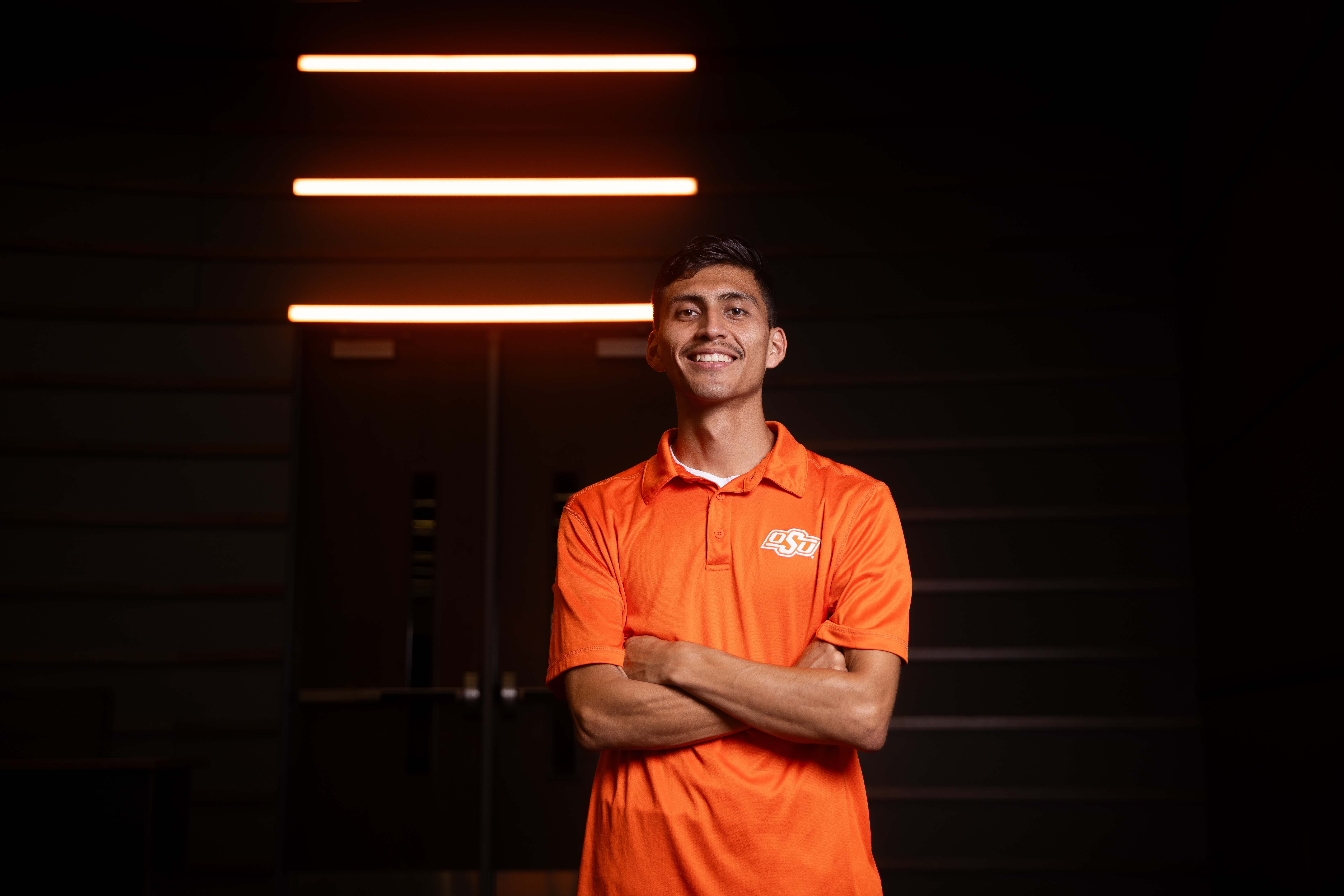 Male student wearing orange polo with crossed arms and smiling.