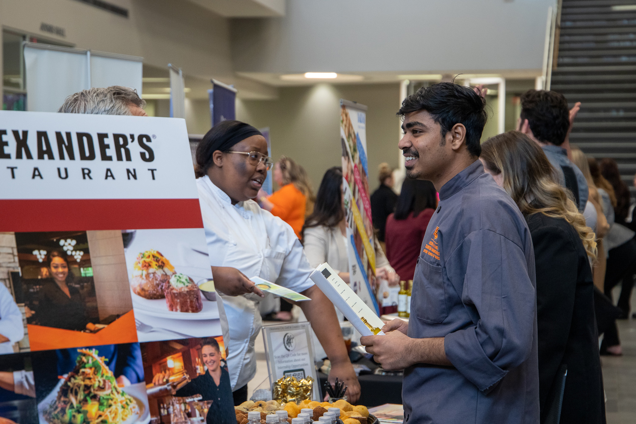 Male student wearing a chef's coat speaking to recruiters at a booth.