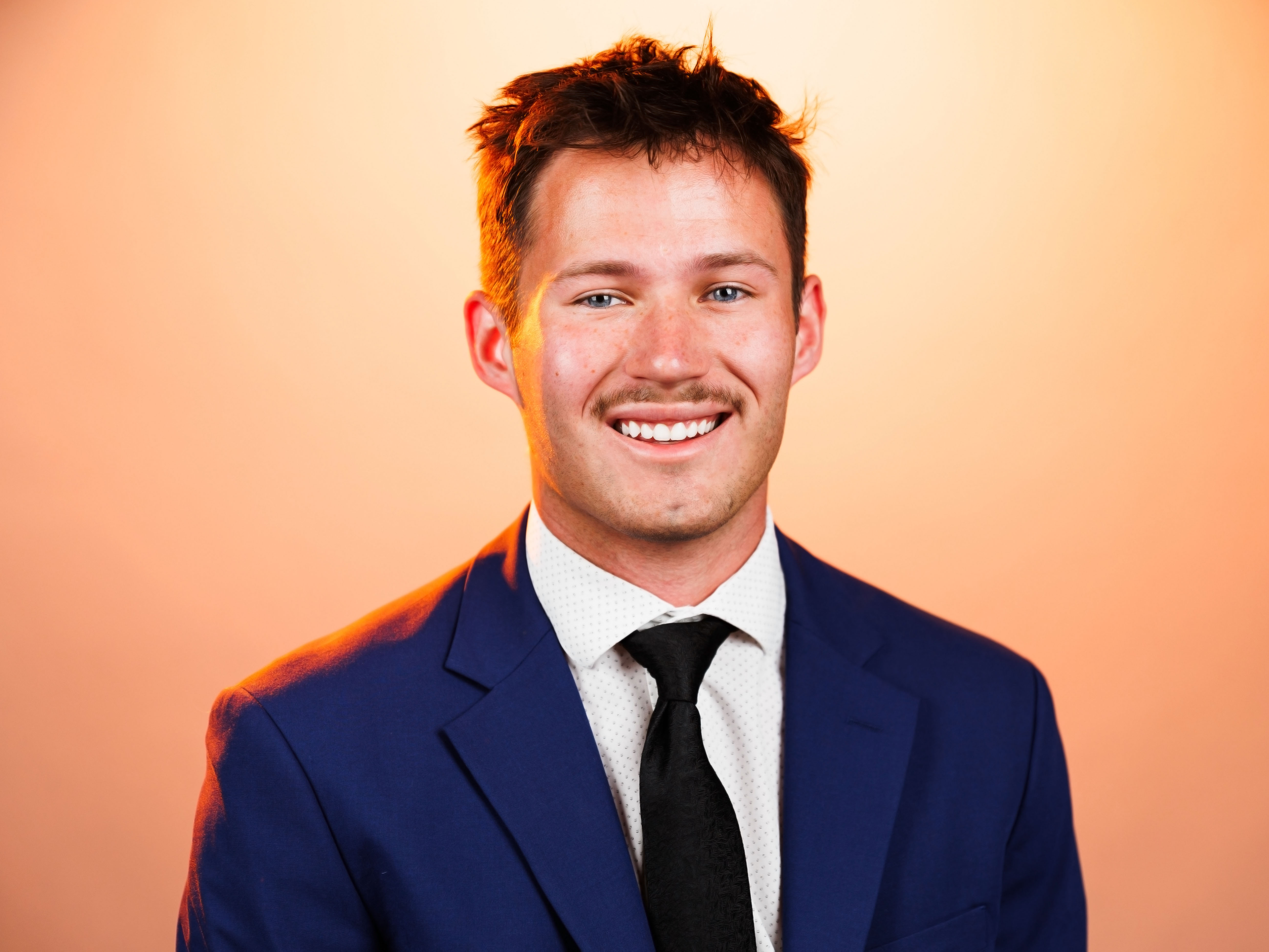 Headshot of Adam Dutka with an orange background. Adam is wearing a suit.