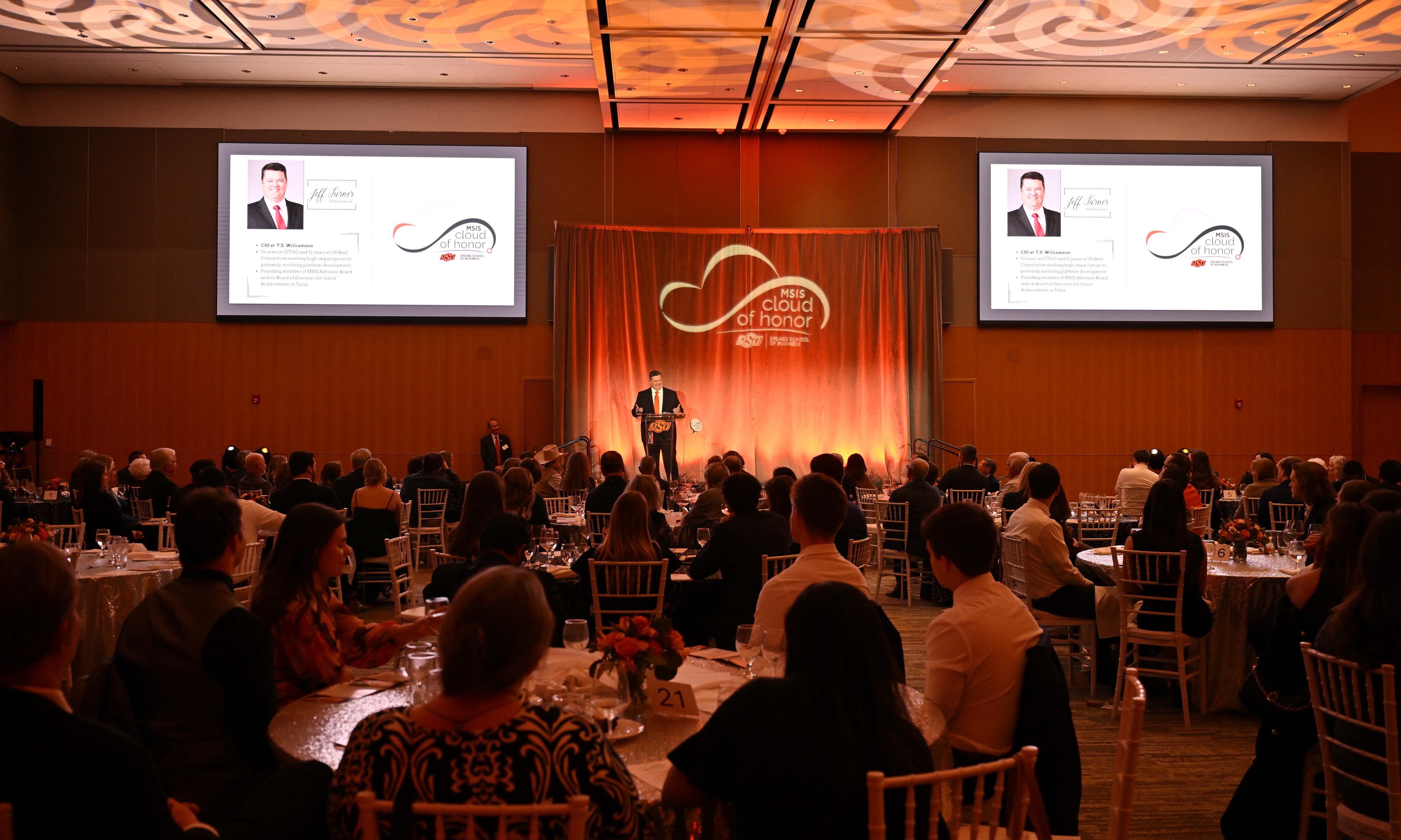 Male alumni speaking from the stage at the MSIS Cloud of Honor celebration in the background. Foreground of full tables listening.