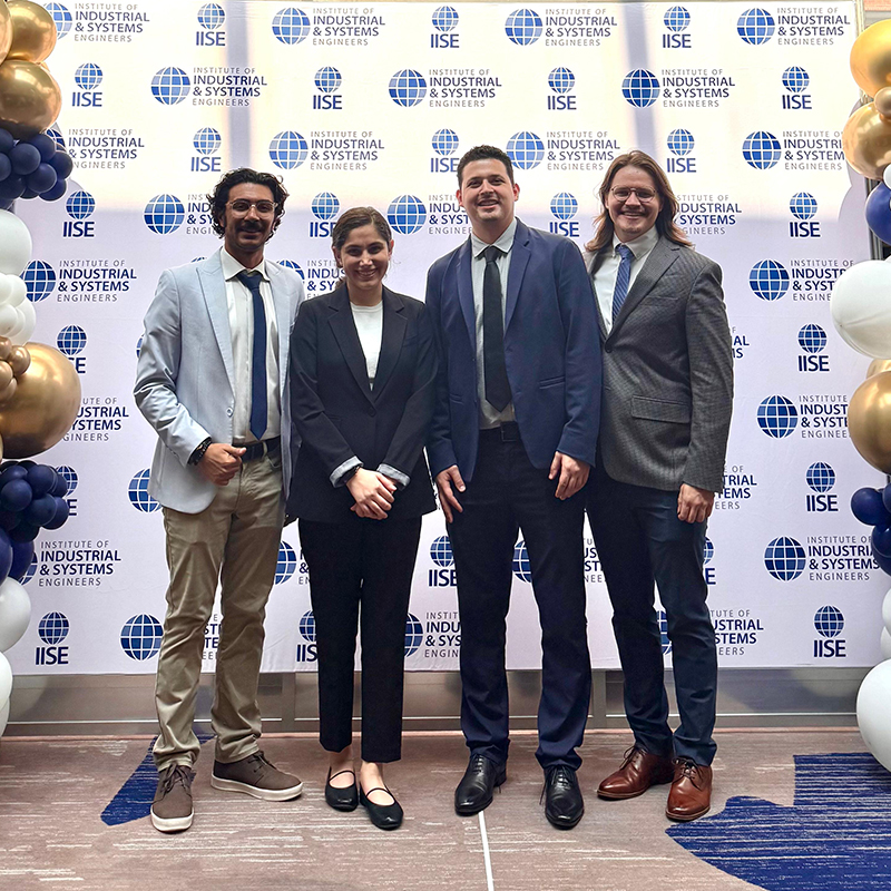 The OSU MS BAnDS team of Edgar Castillo, Bennett Frohock, Mahla Hosseini and Hamed Hemmati pose in front of an IISE banner.