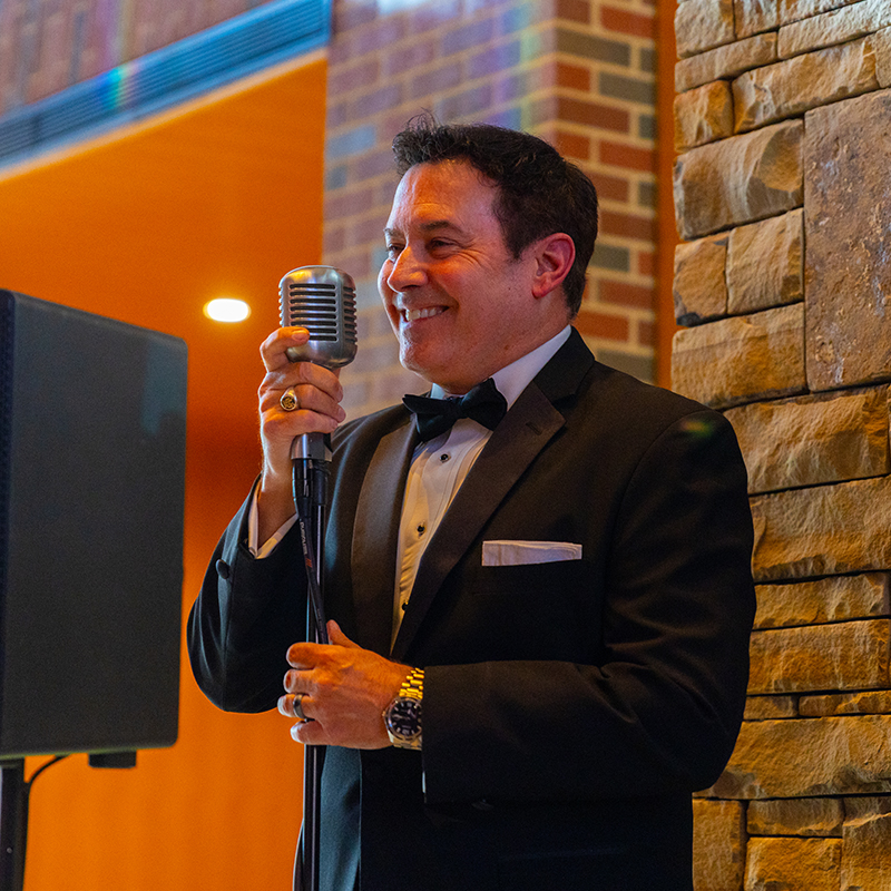 A singer in a tuxedo smiles while singing into an old-timey microphone.
