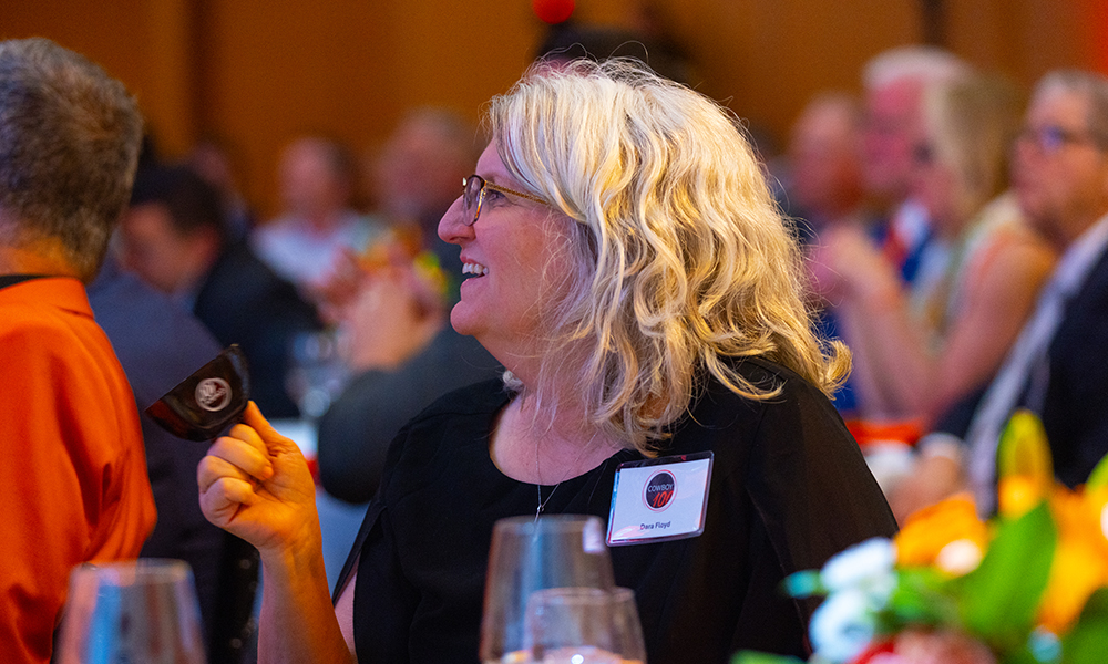 A woman rings a Cowboy100 cowbell noisemaker while smiling at her table.